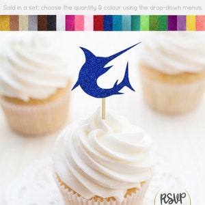 Puede incluir: Topper de cupcake de marlin azul con purpurina. El topper está en un cupcake blanco con glaseado blanco. El cupcake está en un encaje blanco. El topper está hecho de cartulina con purpurina y se vende en un juego.  "RSVP Party Decor" está impreso en una pegatina circular dorada.