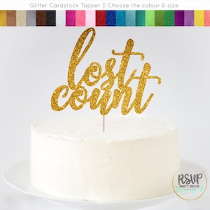 May include: Gold glitter cardstock cake topper with the words "lost count" in a cursive font. The topper is on top of a white cake.