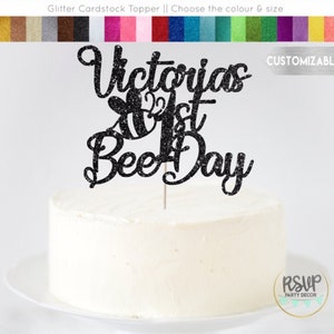 May include: Black glitter cardstock cake topper with the text "Victoria's 1st Bee Day" and a small bee illustration. Customizable.