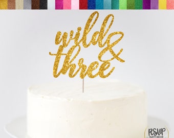 以下が含まれることがあります： 白いフロストケーキの上に「wild & three」と書かれた金色のグリッターケーキトッパー。トッパーは細い銀色の棒の上にあります。ケーキは白いお皿の上にあります。「Glitter Cardstock Topper || Choose the colour & size」という文字が上部にあります。