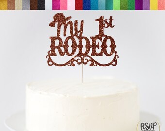 May include: A brown glitter cake topper with the text "My 1st Rodeo" and a cowboy hat on top. The topper is on a white cake.