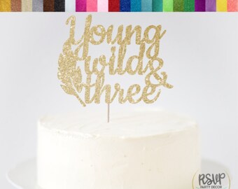 以下が含まれることがあります： ゴールドのグリッターカードストックケーキトッパー。テキストは「Young Wild & Three」で、羽根のデザインが施されています。