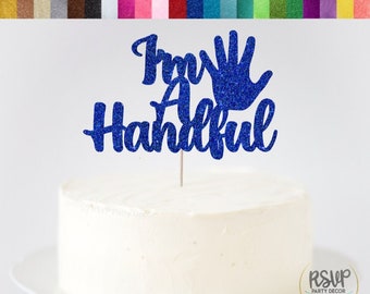 May include: Blue glitter cake topper with the text "I'm a Handful" and a handprint. The topper is on a white cake.