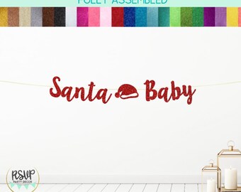 May include: A red glitter banner that reads "Santa Baby" with a Santa hat icon. The banner is strung across a white wall, with two gold lanterns and candles in the foreground. The top of the image shows a color palette of glitter paper.