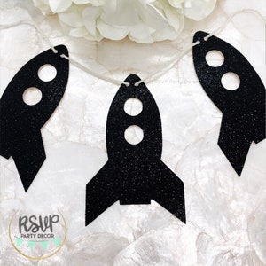 May include: A banner featuring three black glitter rocket ship cutouts strung on twine. Each rocket has two circular windows. The banner is for party decor. The RSVP Party Decor logo is visible in the lower left corner.