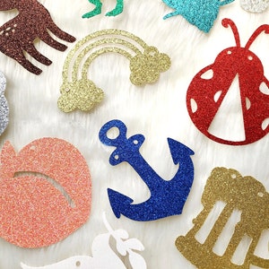 May include: A collection of glitter-covered paper cutouts in various shapes and colours, including a rainbow, an anchor, a ladybug, a peach, a deer, a graduation cap, and a beer mug. The cutouts are arranged on a white fluffy surface.