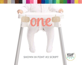 May include: A glittery banner featuring the word one in a coral script font flanked by two gold glitter pumpkins. The banner is attached to a white high chair. A colour palette of various glitter swatches is displayed at the top of the image.