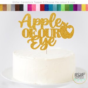 May include: A gold glitter cake topper that reads "Apple of Our Eye" with a heart-shaped apple design. The topper is on a white frosted cake, perfect for a celebration. The image also shows a variety of glitter cardstock colors.