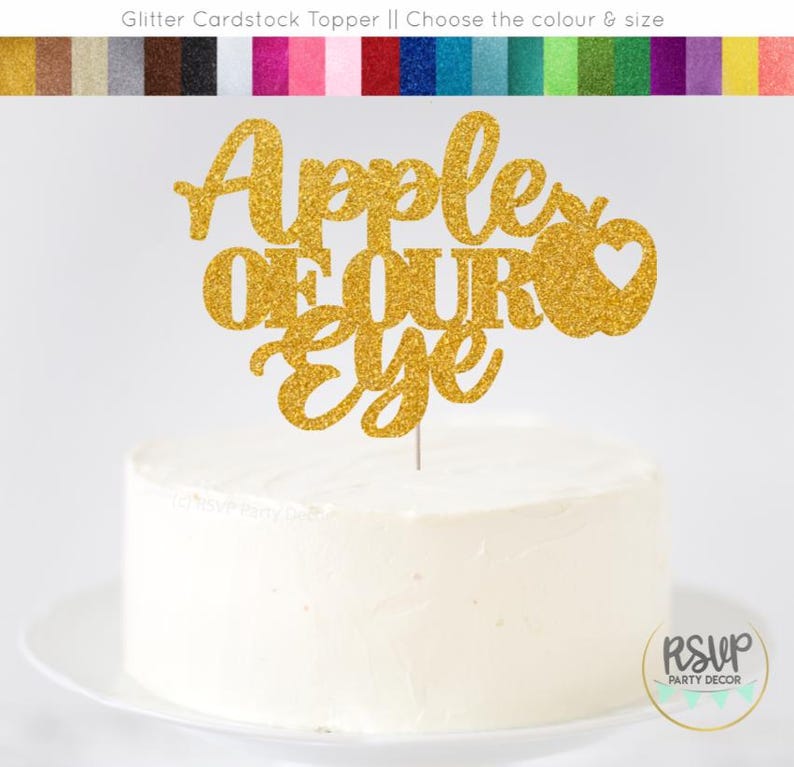 May include: A gold glitter cake topper that reads "Apple of Our Eye" with a heart-shaped apple design. The topper is on a white frosted cake, perfect for a celebration. The image also shows a variety of glitter cardstock colors.
