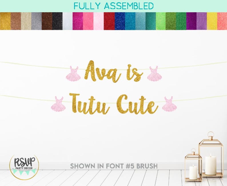 Może przedstawiać: Złota girlanda z błyszczącego papieru z napisem "Ava is Tutu Cute" i wycięciami baletnic z r&oacute;żowego brokatu. Girlanda jest przewleczona przez białą sznur&oacute;wkę i wisi na białej ścianie.