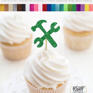 May include: Green glitter cupcake topper with a hammer and wrench design. The topper is on a white cupcake with white frosting.