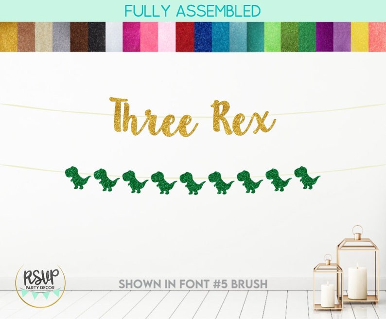May include: A gold glitter banner with the text "Three Rex" and green glitter dinosaur cutouts. The banner is strung on a white string and is shown against a white wall.