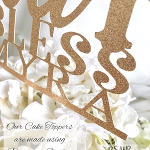 May include: Gold glitter cake topper with the words "Our Bless Myra" on it. The topper is sitting on a white cake with white flowers. The text "Our Cake Toppers are made using Premium Quality Glitter Cardstock" is visible below the cake topper. The text "RSUP Parties & Events" is visible in the bottom right corner of the image.