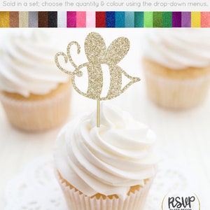 May include: Gold glitter bee cupcake topper. The bee has a striped body and is on a wooden stick. The topper is on a white cupcake with white frosting.