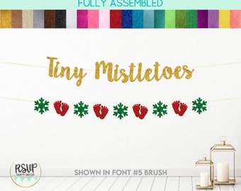May include: Gold glitter banner with the text "Tiny Mistletoes" and alternating red footprints and green snowflakes. The banner is fully assembled and ready to hang.