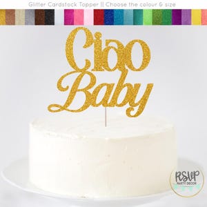 K&ouml;nnte beinhalten: Eine wei&szlig; glasierte Torte mit einem goldglitzernden "Ciao Baby" Tortenaufsatz. Der Aufsatz ist an einem d&uuml;nnen, silbernen Stab befestigt. Die Torte steht auf einem wei&szlig;en Teller. Oben im Bild ist eine Farbkarte aus Glitzerkarton zu sehen.