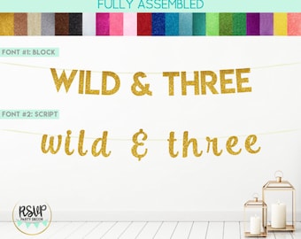 Puede incluir: Bandera de brillo dorado con el texto "WILD & THREE" en letras de bloque y "wild & three" en letras de escritura. La bandera está enhebrada en una cuerda blanca y está colgada en una pared blanca.