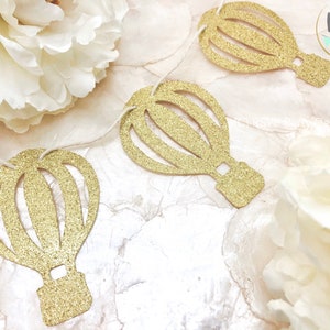 May include: Gold glitter hot air balloon cutouts strung on a white string. The cutouts are arranged on a white and cream-colored surface with white flowers. The balloons are for party decor.