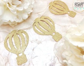 Puede incluir: Recortes de globos aerostáticos dorados con purpurina ensartados en una cuerda blanca. Los recortes están dispuestos sobre una superficie blanca y crema con flores blancas. Los globos son para la decoración de fiestas.