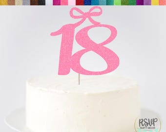 May include: Pink glitter cardstock cake topper with a bow and the number 18. The topper is on a white cake.
