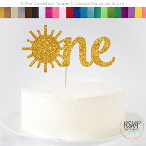 Op de afbeelding: Gouden glitter-taarttopper van karton met een zon en het woord "one" in een cursief schrift. De topper is op een witte taart.