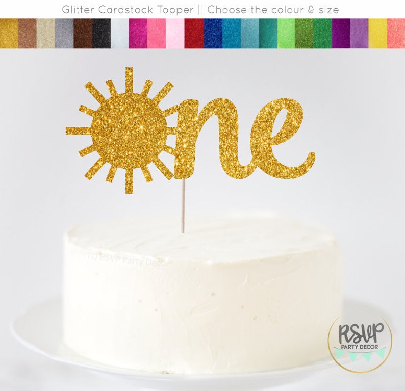 Op de afbeelding: Gouden glitter-taarttopper van karton met een zon en het woord "one" in een cursief schrift. De topper is op een witte taart.