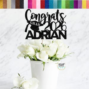 May include: A black glitter cake topper with the words "Congrats Grad 2026 ADRIAN" and a graduation cap. The topper is placed in a white vase filled with white roses. A variety of glitter paper colours are displayed in the background.