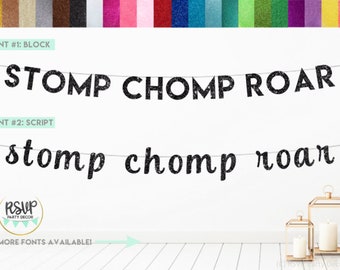 May include: Black glitter banner with the words "STOMP CHOMP ROAR" in block and script fonts. The banner is hanging on a white wall with two gold lanterns in the foreground.