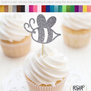 May include: Silver glitter bee cupcake topper. The bee has black and white stripes. The topper is on a cupcake with white frosting. The cupcake is on a white doily. The text "RSUP PARTY DECOR" is in the lower right corner of the image.