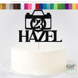 May include: Black glitter cardstock cake topper with a camera design and the number 23 inside the camera lens. The name Hazel is written below the camera.