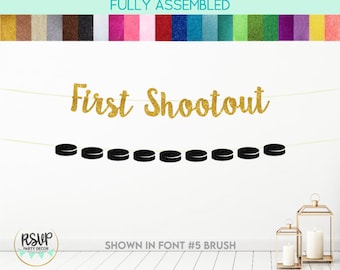 May include: Gold glitter "First Shootout" banner with black hockey puck cutouts. The banner is strung on a white string and is shown against a white wall.