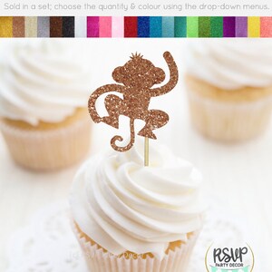 K&ouml;nnte beinhalten: Goldener Glitzer-Affen-Cupcake-Topper. Der Affe sitzt mit erhobenen Armen und seinem Schwanz um sein Bein gewickelt. Der Topper befindet sich auf einem wei&szlig;en Cupcake mit wei&szlig;em Frosting.