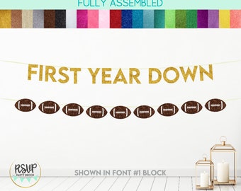 May include: A gold glitter banner that reads "FIRST YEAR DOWN" with brown football decorations. The banner is fully assembled and shown in font #1 block. RSVP Party Decor is in the bottom left corner.