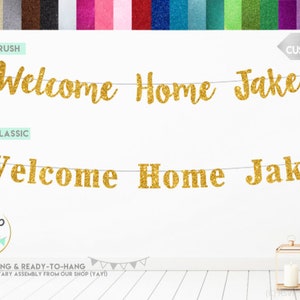Pode incluir: Faixa de boas-vindas para casa de glitter dourado com o nome Jake. A faixa &eacute; pr&eacute;-enfiada e pronta para pendurar. A faixa &eacute; mostrada pendurada em uma parede branca com duas lanternas douradas em primeiro plano.