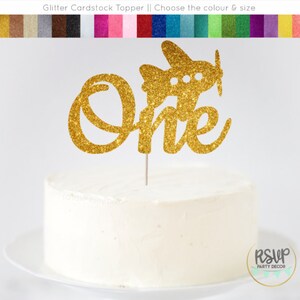 May include: Gold glitter cardstock cake topper with the word "one" in a cursive font and a small airplane on top. The topper is on a white cake.