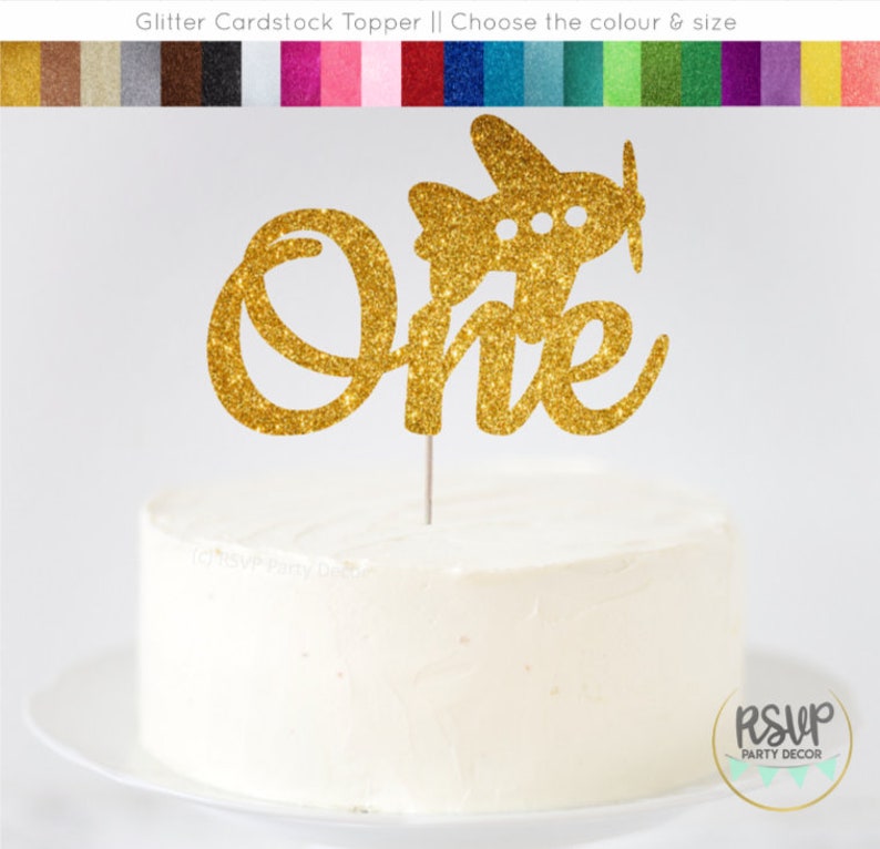 May include: Gold glitter cardstock cake topper with the word "one" in a cursive font and a small airplane on top. The topper is on a white cake.