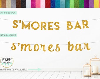 May include: Gold glitter S'mores Bar banner with two different font styles. The banner is hanging on a white wall with a wooden floor. The text on the banner reads "S'MORES BAR" in block letters and "s'mores bar" in script letters.