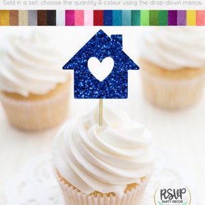 May include: Blue glitter cupcake topper shaped like a house with a white heart inside. The topper is on a wooden stick and is perfect for a housewarming party or a new home celebration.