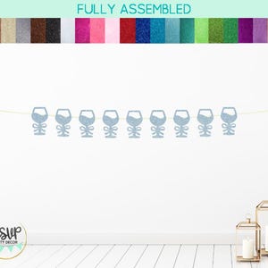 Könnte beinhalten: Ein hellblauer Glitzer-Wein-Glas-Banner mit dem Text "RSVP Party Decor" in einem weißen Kreis mit grünem Rand. Der Banner ist an einer weißen Schnur befestigt und hängt an einer weißen Wand.