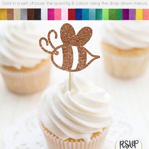 May include: Gold glitter cupcake topper shaped like a bee. The bee has white stripes and a brown body. The topper is on a cupcake with white frosting. The cupcake is on a white doily. The text "RSUP PARTY DECOR" is in the lower right corner of the image.