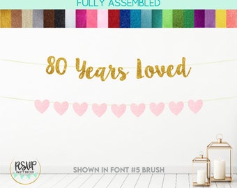 Puede incluir: Pancarta dorada con purpurina con el texto "80 Years Loved" y una hilera de corazones rosas con purpurina. La pancarta se muestra en la fuente #5 brush.