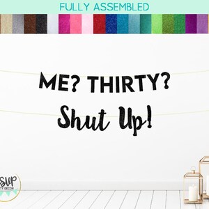 Puede incluir: Pancarta negra de purpurina con el texto "ME? THIRTY? SHUT UP!" colgada en una pared blanca. La pancarta est&aacute; hecha de letras individuales y est&aacute; enhebrada con una cuerda dorada.