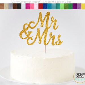 May include: Gold glitter cardstock cake topper with the words "Mr & Mrs" in a cursive font. The topper is on top of a white cake.