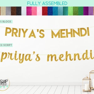 Puede incluir: Pancarta de purpurina dorada con el texto "PRIYA'S MEHNDI" en letras de bloque y "priya's mehndi" en letras de escritura. La pancarta está enhebrada en una cuerda blanca y está colgada en una pared blanca.