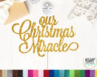 May include: A gold glitter cake topper with the words "Our Christmas Miracle" in a cursive font. The topper is set against a white wooden background with a gift box and decorative elements.