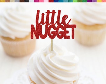 Könnte beinhalten: Ein roter Glitzer-Cupcake-Topper mit dem Text "little NUGGET" auf einem weißen Cupcake mit weißem Frosting. Der Topper ist auf einem Holzstiel.