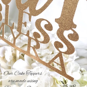 Op de afbeelding: Gouden glittertaarttopper met de woorden "Our Bless Myra" erop. De topper staat op een witte taart met witte bloemen. De tekst "Our Cake Toppers are made using Premium Quality Glitter Cardstock" staat onder de taarttopper. De tekst "RSUP Parties & Events" staat in de rechterbenedenhoek van de afbeelding.