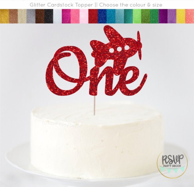 May include: Red glitter cardstock cake topper with the word "one" and a small airplane. The topper is on a white cake.