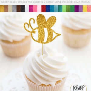May include: Gold glitter bee cupcake topper. The bee has black and white stripes. The topper is on a white cupcake with white frosting. The cupcake is on a white doily. The text "RSUP PARTY DECOR" is in the lower right corner of the image.