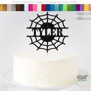May include: Black glitter cardstock cake topper featuring a spiderweb design with the name "TYLER" in the centre. The topper is on a white cake.